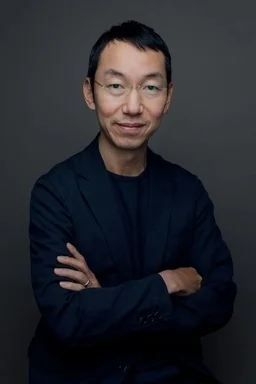 暗い背景を背に、メガネをかけたアジア人男性が腕を組んでこちらを見ているポートレート。