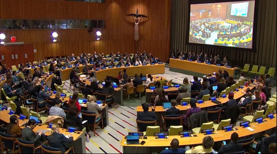 国際会議の会場で、多数の参加者が半円状に配置された席に着席し、正面の巨大スクリーンには会議の様子が映し出されています。活発な議論が展開されている雰囲気です。