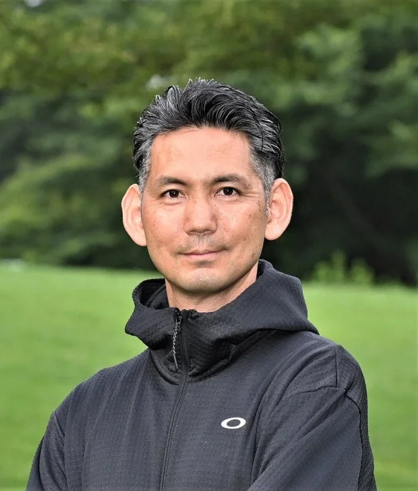 緑豊かな屋外を背景に、グレーの髪をしたアジア人男性が黒いフード付きジャケットを着て立っています