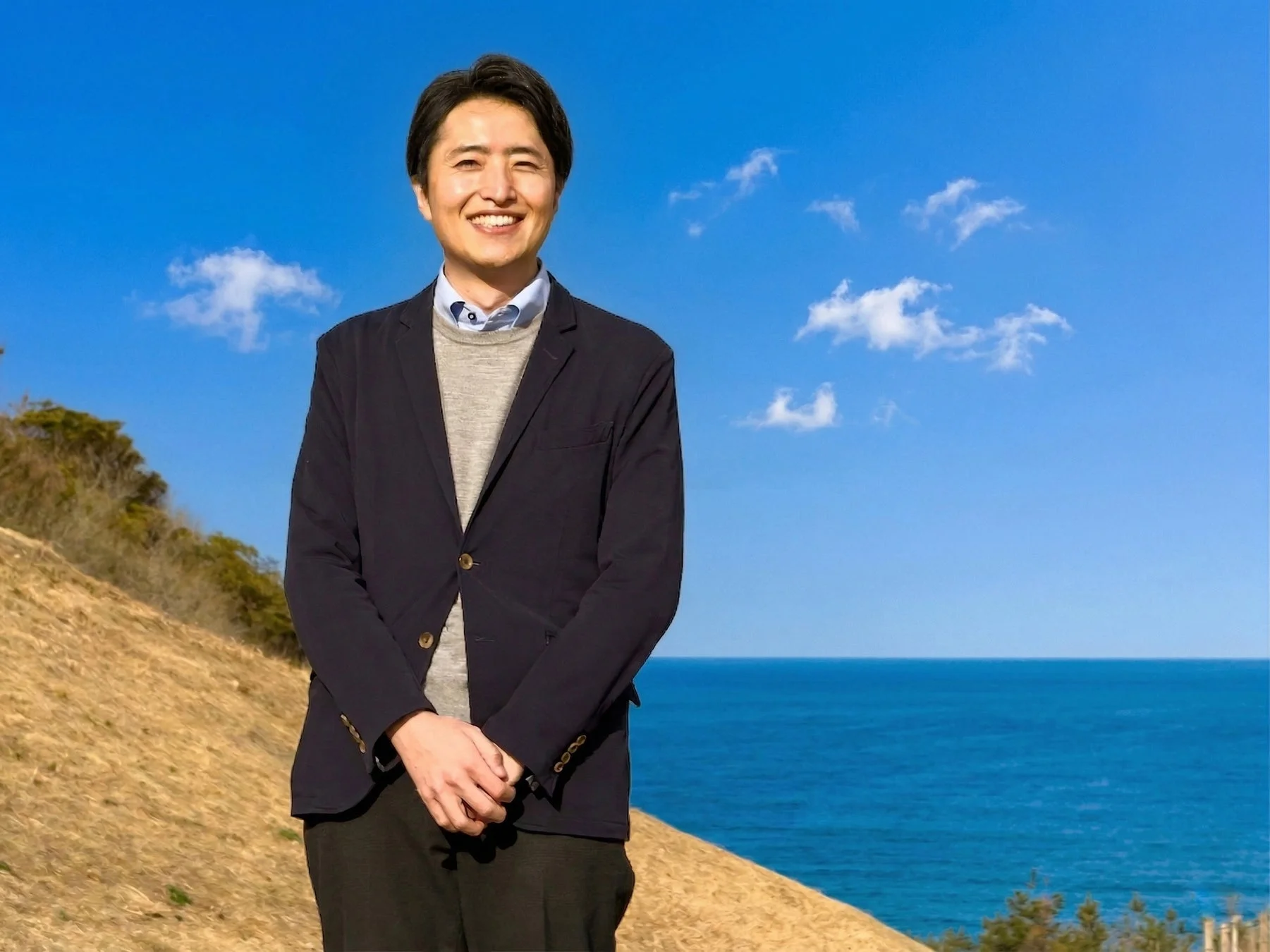 海と丘を背景に笑顔で立つ男性（田口真氏）