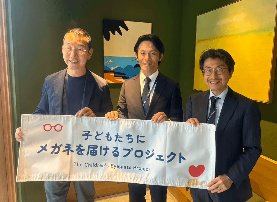 3人の男性が「子どもたちにメガネを届けるプロジェクト」と書かれたバナーを笑顔で掲げている写真です。