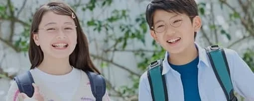 眼鏡をかけた少年と少女がリュックサックを背負い、屋外で楽しそうに笑顔を見せています。