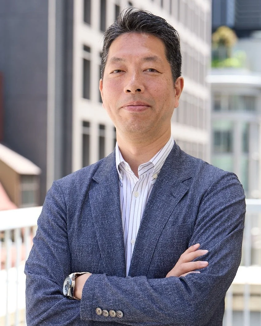 ビジネスカジュアルな服装の日本人男性が、屋外の都市を背景に腕を組んで立っているポートレート写真です。穏やかな表情でカメラを見ています。